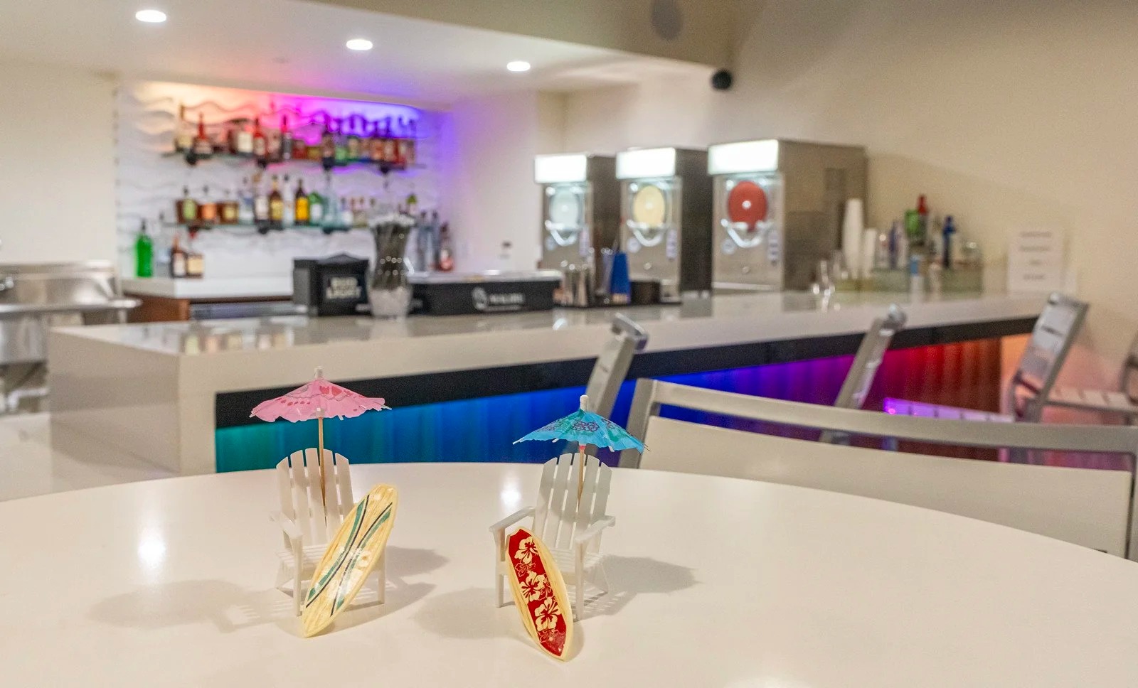 Tropical-themed bar with colorful lights, decorative mini deck chairs, tiny surfboards, and cocktail umbrellas, with a backdrop of alcohol bottles and slush machines on the counter.