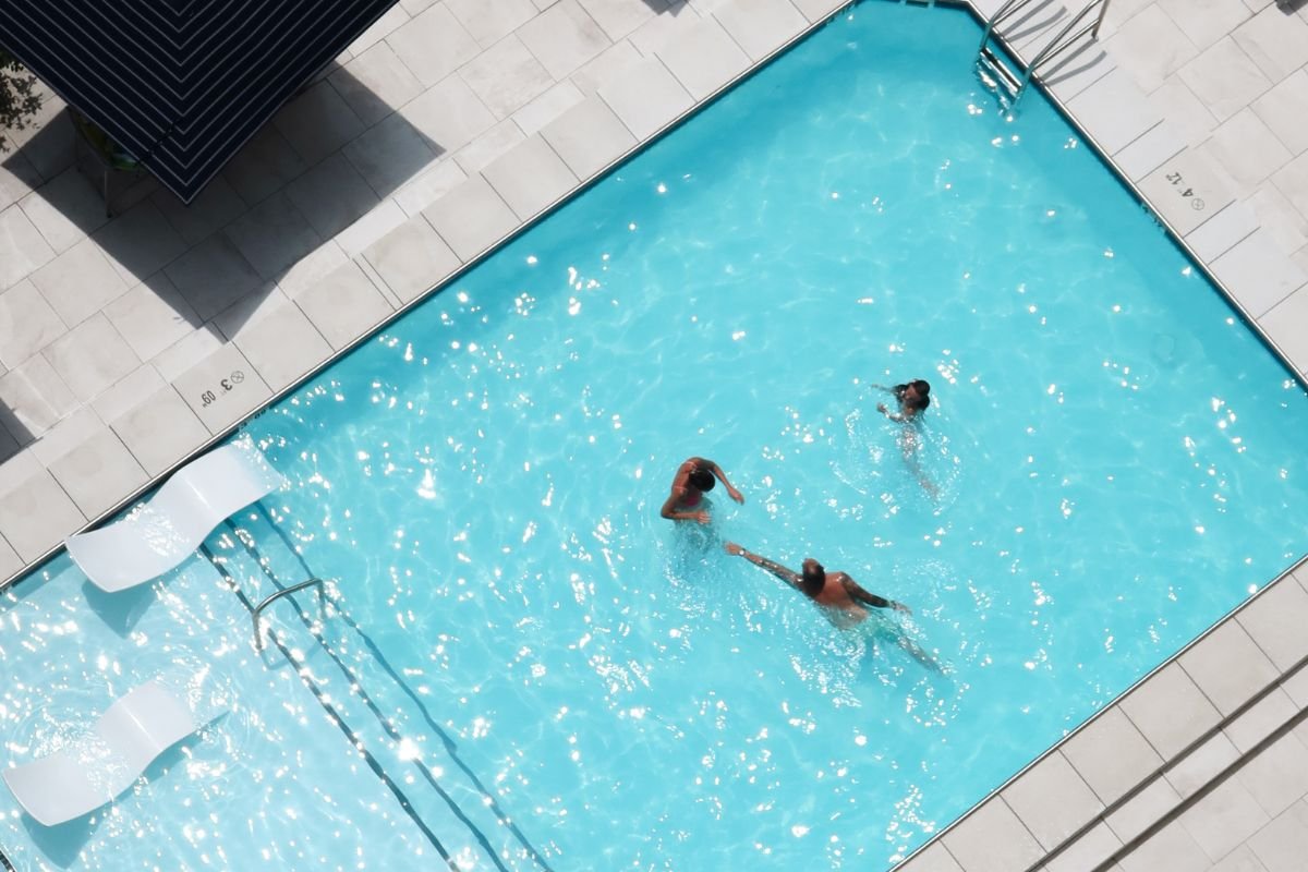 Aerial view of three people swimming in a bright blue swimming pool. Two white lounge chairs are submerged at the pool edge. The pool is surrounded by a light-colored tiled deck.