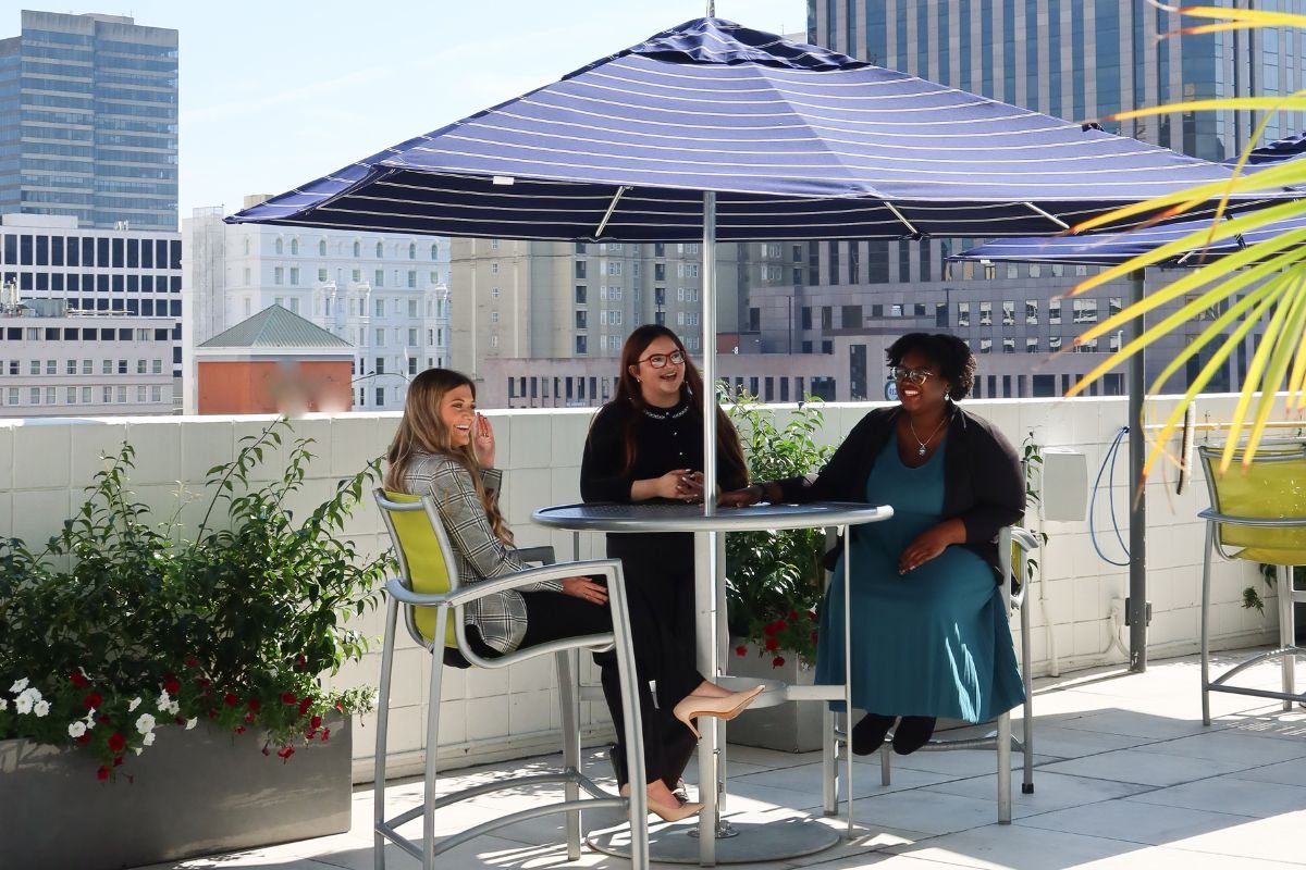 Three women sitting at a patio table under a blue umbrella, laughing and conversing, with an urban skyline in the background.