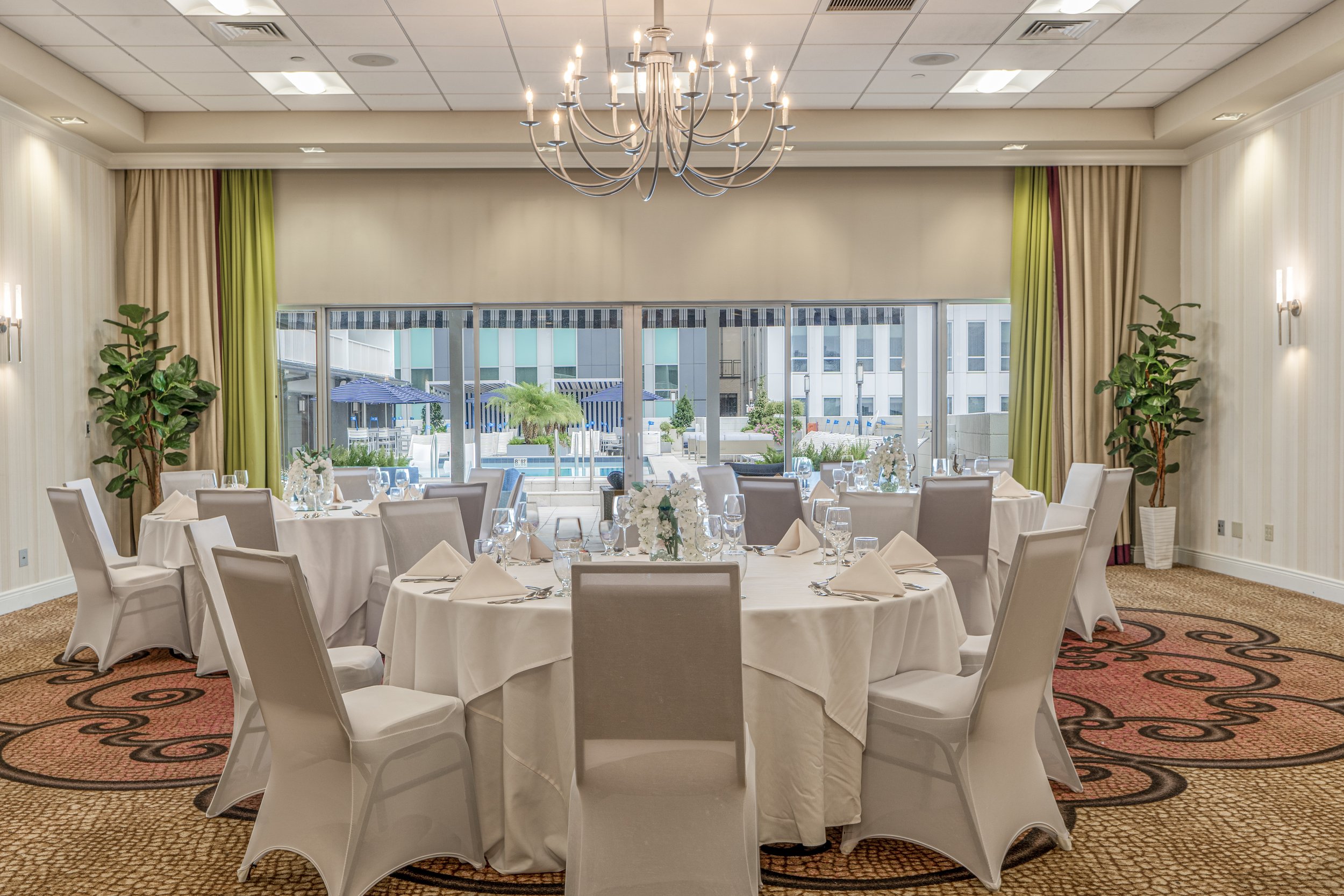 Elegant banquet hall with round tables, white tablecloths, and formal place settings. A chandelier hangs from the ceiling. Large windows provide a view of a patio area with greenery. The room has a neutral color scheme with decorative lighting and po