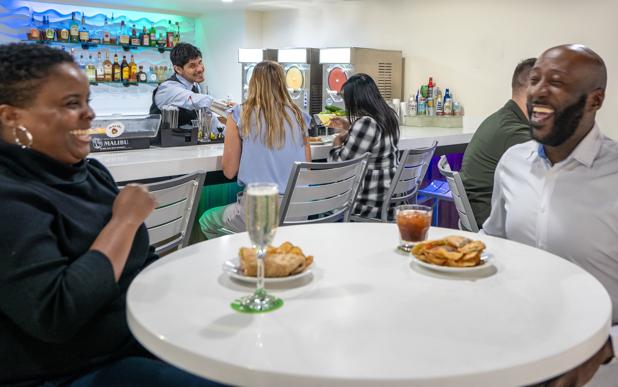People enjoying food and drinks at a bar, with a smiling bartender in the background.