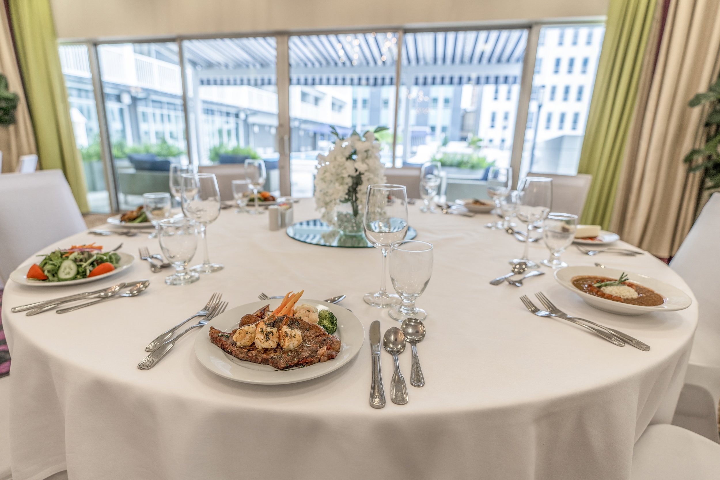 Elegant dining table set with white tablecloth, empty glasses, cutlery, and plates of gourmet food; centerpiece with white flowers; bright indoor setting overlooking a terrace.