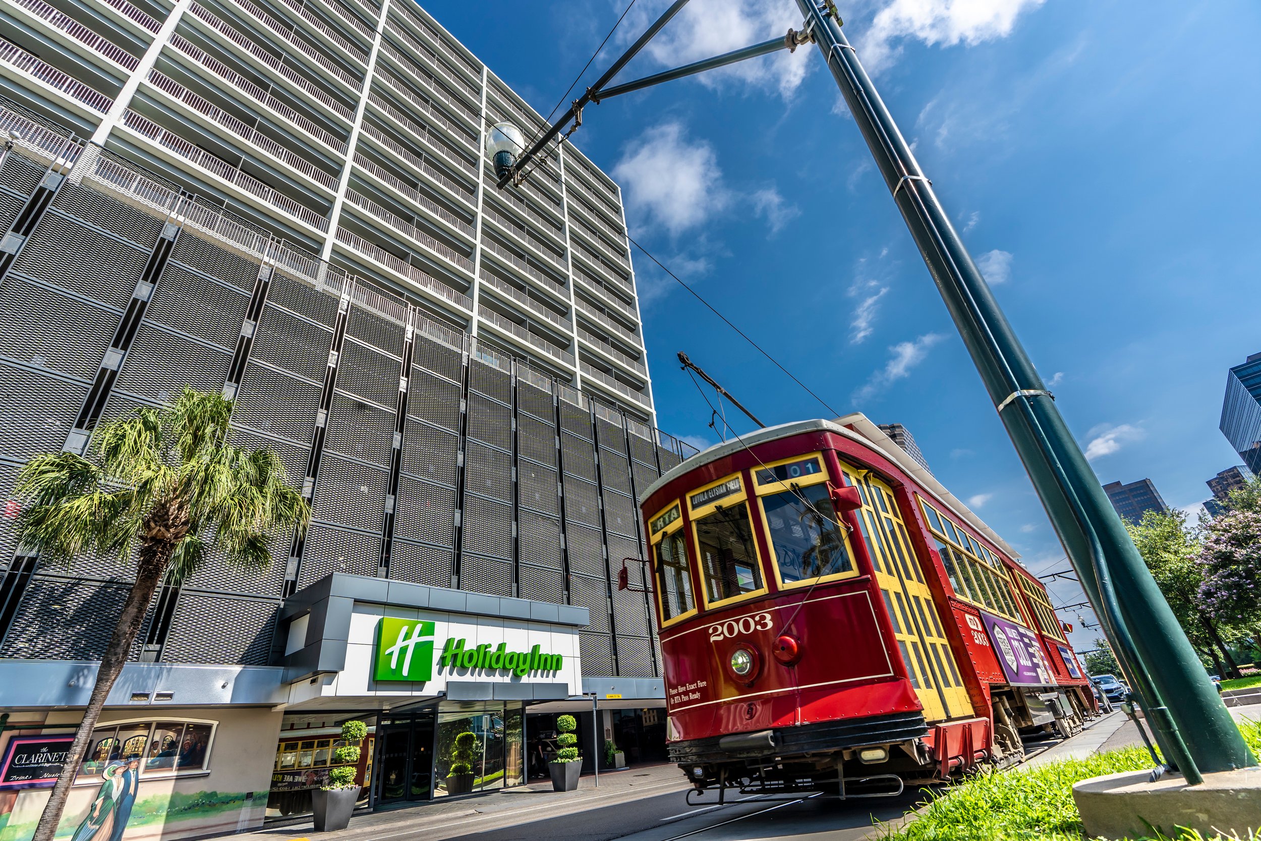 Red streetcar passing by a Holiday Inn hotel with a high-rise building, palm trees, and clear blue sky in New Orleans.