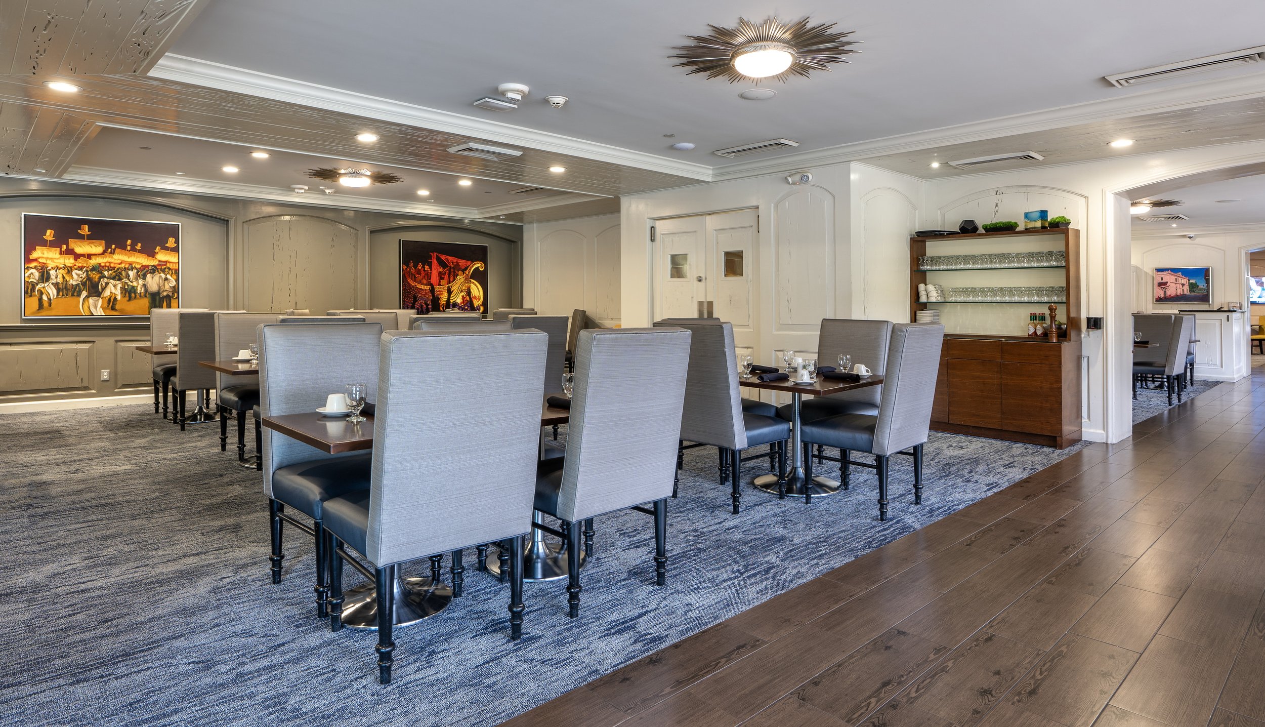 Interior of a modern restaurant with gray high-back chairs and wooden tables, a blue carpet, and hardwood floors. The walls feature colorful artwork, and there's a sideboard with glasses and cups. The lighting is contemporary.