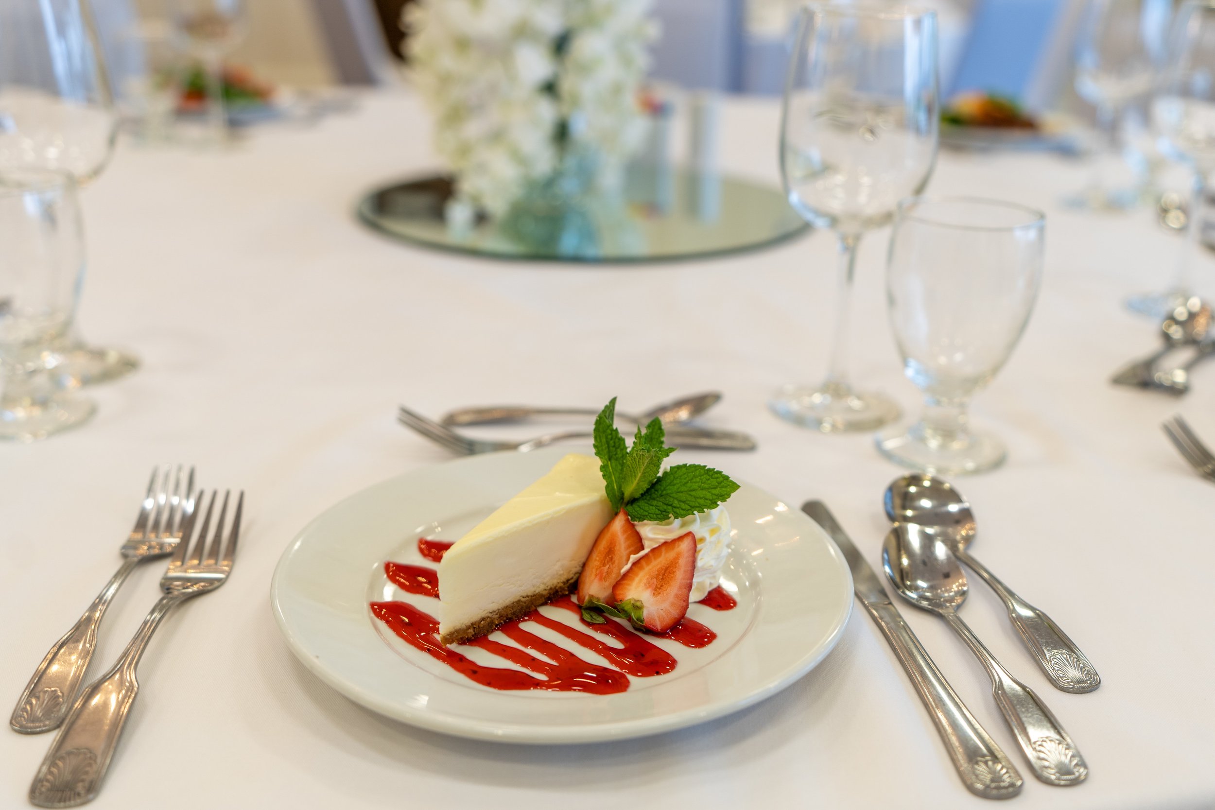 A slice of cheesecake with strawberries and mint garnish on a white plate, surrounded by elegant tableware on a formal dining table.