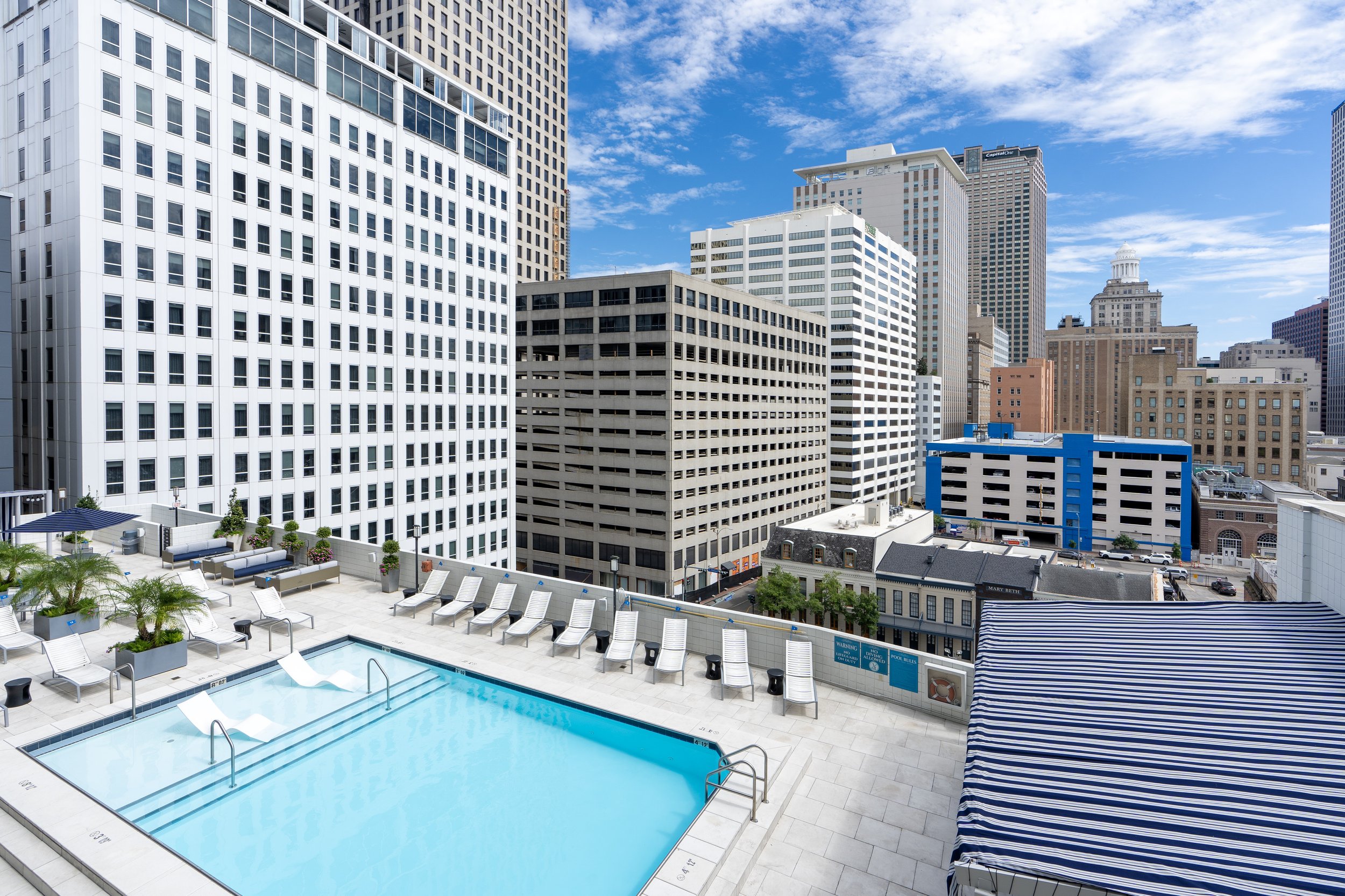 Rooftop swimming pool with lounge chairs, surrounded by tall buildings in an urban setting, under a blue sky.